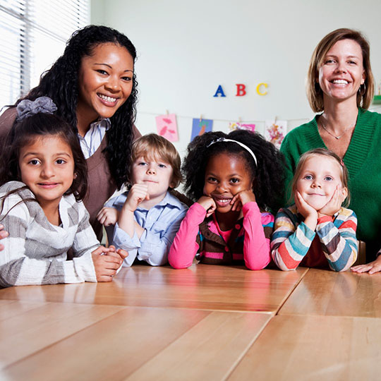 two preschool teachers smiling with 4 children at a classroom table